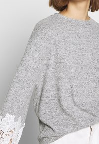 Ljusgrå tröja med långa ärmar som har delikata vita spetsdetaljer vid manschetterna. Mjuk textur, enkel rund halsringning.