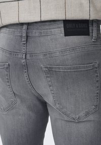 Blugi din denim gri cu o croială de tip mid-rise, având două buzunare la spate și o etichetă din piele inscripționată "ONLY & SONS" la talie.