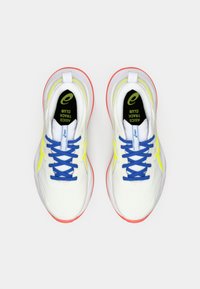 Baskets de sport blanches avec des lacets bleus, des accents jaunes et une semelle rouge. Présentent un dessus texturé et un col rembourré pour le confort.