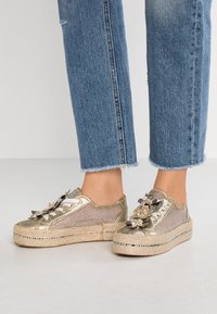 Zapatillas deportivas de plataforma doradas metálicas con acentos florales, textura tejida y suela de estilo yute. Llevadas con jeans de mezclilla azul con dobladillo.