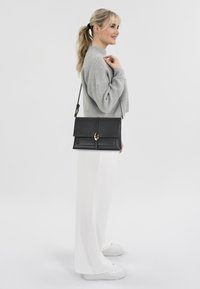 Borsa a tracolla nera in pelle con pattina pieghevole, accessori in oro e tracolla removibile, abbinata a un maglione grigio e pantaloni bianchi.