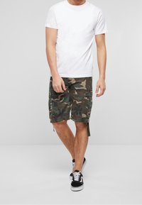 Bílá tričko s krátkým rukávem v kombinaci s zelenými maskáčovými cargo šortkami. Šortky mají více kapes a nastavitelné boční popruhy. Černé tenisky.