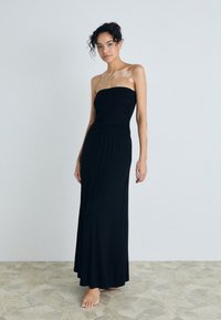Svart stroppeløs maxikjole i myk stoff, tettsittende bodice, flytende skjørt og rynket midje detalj; modellen står på teksturert gulv.