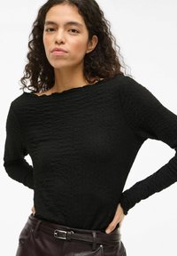 Czarna bluzka z długim rękawem o fakturowanej powierzchni, z dekoltem w łódkę i lekko postrzępionymi krawędziami. Noszona z ciemnymi bordowymi spodniami i srebrnym paskiem.