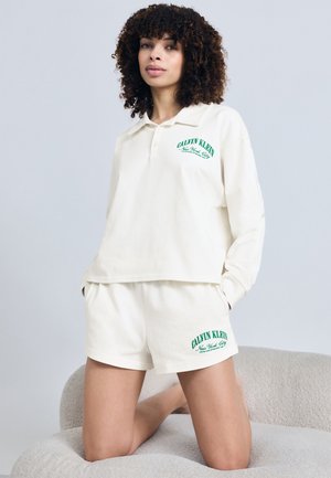 Femeie cu păr creț purtând un polo alb cu mâneci lungi și pantaloni scurți asortați cu logo-uri verzi „Calvin Klein New York City”, genuflexată pe un scaun cu textură moale.