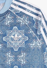 Tela azul de Adidas con intrincados patrones geométricos y florales en blanco y rosa, y las características tres franjas blancas en el hombro.