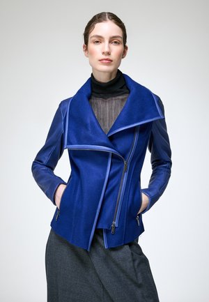 Femme portant une veste bleue avec fermeture éclair asymétrique et col, un col roulé noir et une jupe gris foncé, debout les mains dans les poches.