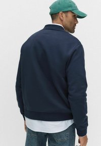 Mies, który ma na sobie granatowy sweter, jasnoniebieską koszulę, dżinsy i zieloną czapkę z daszkiem, odwrócony plecami i patrzący w bok na jednolitym tle.