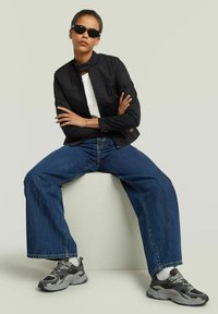 Giacca nera cropped con colletto a stand, abbinata a jeans a gamba larga blu e sneakers grigie. Modella che indossa occhiali da sole scuri.