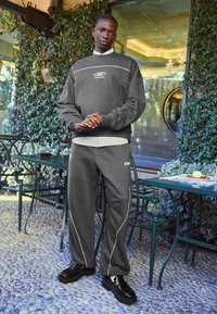 Siva pulover s vezenim logotipom, v kompletu s sivimi trenirkami, ki imajo bele akcentne detajle. Črne loafers čevlje dopolnjujejo izgled.