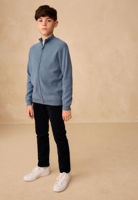 Blå trøje med lynlås lavet af strikket stof, kombineret med sorte manchesterbukser og hvide sneakers, stående på en neutral baggrund.