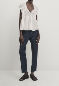 Blouse blanche à manches courtes avec un décolleté en V profond, détails froncés à la taille, associée à un jean droit bleu marine et des sandales noires.