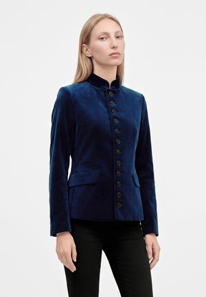 Femme aux cheveux blonds et raides portant une veste en velours bleu foncé cintrée avec un col montant et des boutons noirs, associée à un pantalon noir.