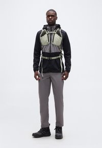 Άνδρας που στέκεται φορώντας μαύρο και γκρι εξωτερικό μπουφάν, γκρι παντελόνι, μαύρα ορειβατικά μποτάκια και ανοιχτό πράσινο σακίδιο πλάτης μπροστά σε απλό λευκό φόντο.