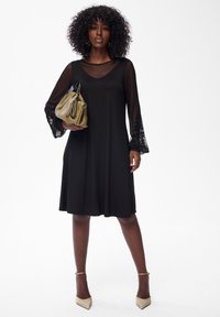 Frau in einem knielangen schwarzen Kleid mit durchsichtigen floralen Ärmeln, die eine olivgrüne Handtasche hält und in beigen High Heels vor einem weißen Hintergrund steht.
