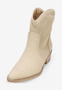 Botín de ante beige hasta el tobillo con diseño perforado, punta afilada y tacón apilado. Presenta una textura suave y un estilo casual.