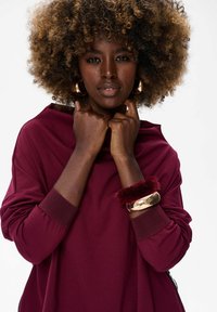 Vrouw met krullend haar, gekleed in een bordeauxrood top, gouden hoepel oorbellen en een gouden bordeauxrood bont armband, die de kraag van haar top vasthoudt.