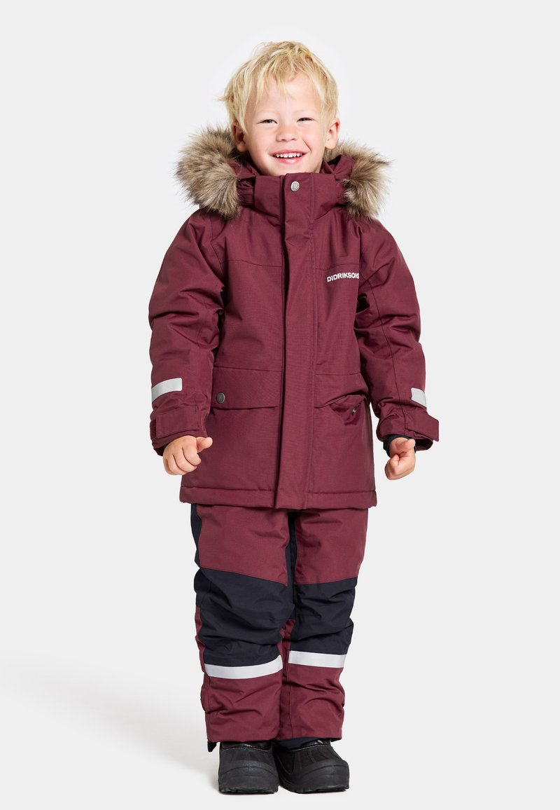 Burgundyröd vinterjacka med falsk päls på huvan, i kombination med marinblå snöbyxor med reflexstriper. Tillverkad av texturerat, isolerat tyg.