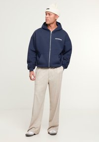 Navy zip-up hoodie with white logo, paired with beige wide-leg pants and gray shoes, finished with a cream beanie hat. Smooth textures.