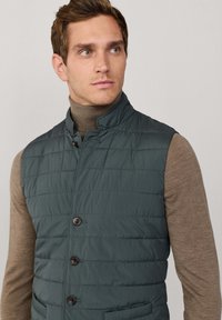 Gepolsterte dunkeltealfarbene Weste mit horizontalem Steppmuster, Stehkragen und braunen Knöpfen. Wird über einem hellbraunen Rollkragenpullover getragen.