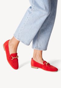 Červené semišové loafers s zlatým reťazovým akcentom, nízkym podpätkom a zaoblenou špičkou, spárované s svetlomodrými skrátenými džínsami.
