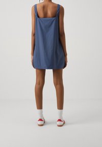 Modré šaty s čtvercovým výstřihem a širokými ramínky, vyrobené z lehkého materiálu. Nosit s bílými ponožkami a botami s červenými akcenty.