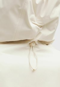 Gros plan d'une capuche de veste beige avec cordon réglable et toggle en plastique rond pour ajuster la coupe.