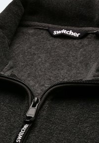 Schwarze Fleece-Jacke mit Reißverschluss und hohem Kragen, YKK-Reißverschluss und "Switcher"-Markierung im Inneren des Etiketts. Weicher, strukturierter Stoff.