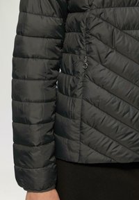 Schwarze Pufferjacke mit einem Chevron-Muster, ausgestattet mit einer Reißverschlusstasche und geripptem Bündchen. Hergestellt aus glänzendem, leichtem Material.