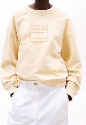 Personne portant un sweat-shirt beige avec le texte "Hilfiger New York 1985" et un graphisme, associé à un pantalon blanc et les mains dans les poches.