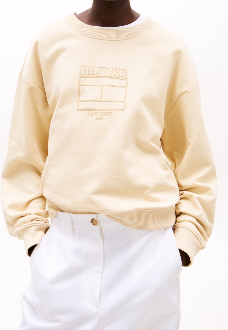 Personne portant un sweat-shirt beige avec le texte "Hilfiger New York 1985" et un graphisme, associé à un pantalon blanc et les mains dans les poches.
