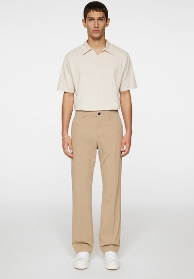 Beige Poloshirt mit kurzen Ärmeln und offenem Kragen, kombiniert mit hellbraunen Hosen. Glatter Stoff und lockere Passform, getragen mit weißen Sneakern.