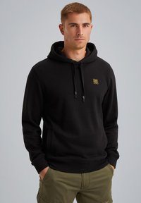 Black hoodie with a drawstring hood, front pocket, and a small yellow logo patch. Material appears soft and textured. Paired with olive cargo pants.