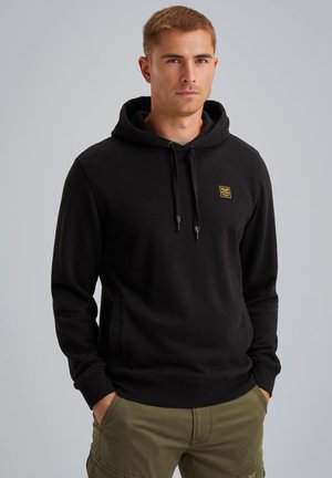 Haut à capuche noir avec un cordon de serrage, poche avant et un petit patch logo jaune. Le matériau semble doux et texturé. Associé à un pantalon cargo olive.