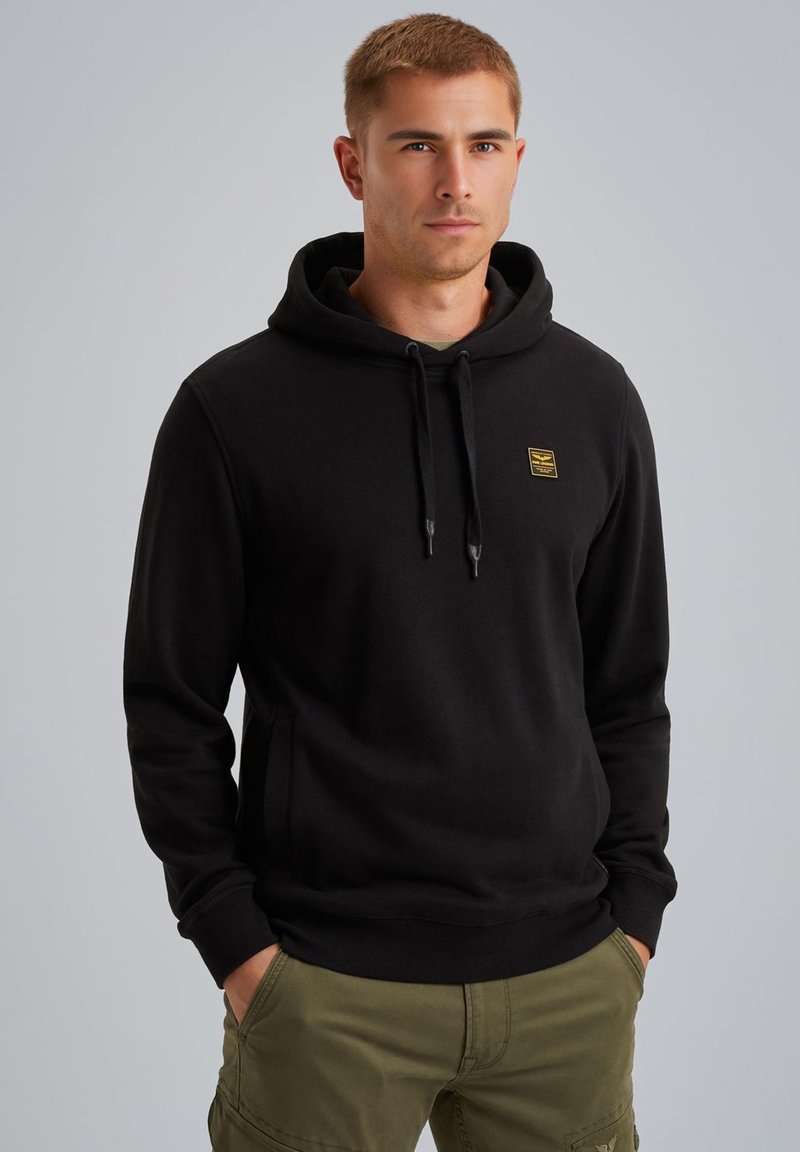 Black hoodie with a drawstring hood, front pocket, and a small yellow logo patch. Material appears soft and textured. Paired with olive cargo pants.