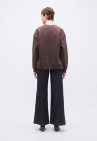 Barna pulóver laza szabással, bordázott mandzsettákkal és kerek nyakkivágással, széles szárú sötét farmernadrággal és barna magassarkú csizmával párosítva.