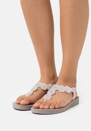 Sandalias de dedo de color rosa claro con acentos circulares adornados con cristales, que cuentan con una suela plana y texturizada de color gris y una correa ajustable.