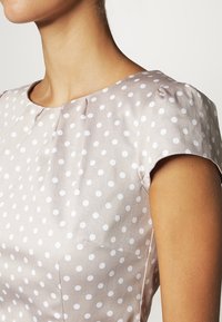 Blusa beige a pois con maniche corte, caratterizzata da pois bianchi, scollatura tonda e una texture di tessuto morbida. Include una tasca laterale.