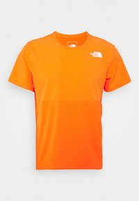 Camiseta naranja de manga corta con una textura suave y paneles en contraste; presenta un pequeño logo blanco en la parte superior izquierda.