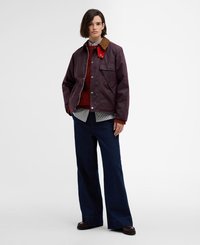 Burgundowa kurtka z brązowym kołnierzem z kordurowej tkaniny, z zatrzaskami i przednią kieszenią, noszona na paskowanym T-shircie i czerwonym swetrze z dzianiny, z szerokimi dżinsami.