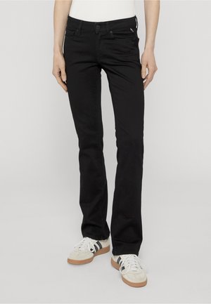 Personne portant un jean noir slim et des baskets blanches avec des bandes noires, debout sur un fond gris clair.