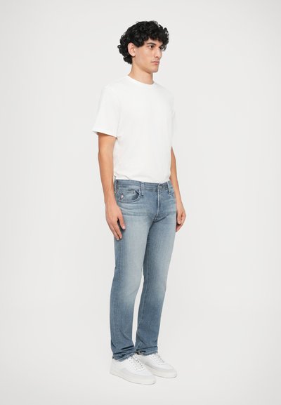 Λευκό μπλουζάκι με στρογγυλή λαιμόκοψη, ανοιχτό μπλε στενές τζιν παντελόνες με ξεθωριασμένο φινίρισμα και λευκά αθλητικά παπούτσια. Στάση με ουδέτερη έκφραση, πλαϊνή άποψη.