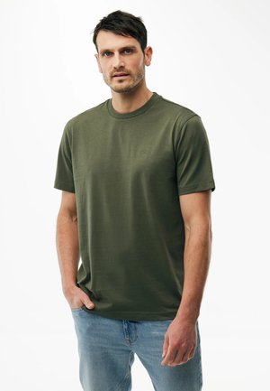 Mann mit kurzen dunklen Haaren trägt ein olivgrünes T-Shirt mit kurzen Ärmeln und Rundhalsausschnitt sowie hellblaue Jeans, steht vor weißem Hintergrund.