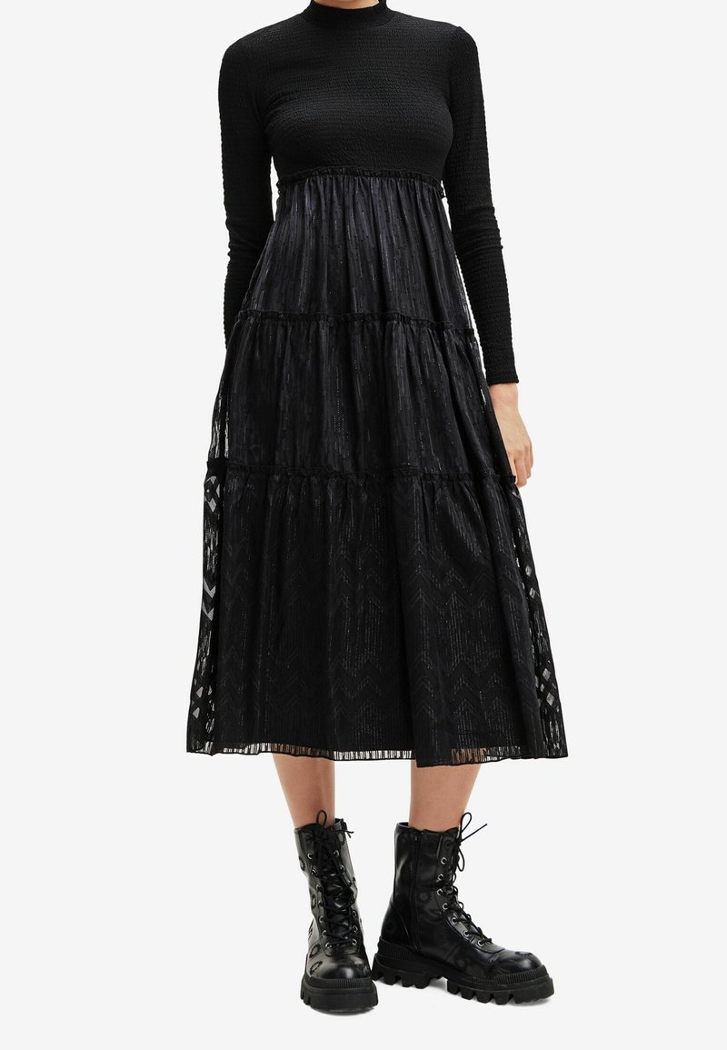 Femme portant un top noir à manches longues et une jupe midi noire transparente à volants avec des bottines de combat noires lacées.