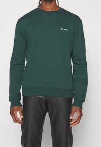 Ciemnozielona bluza z długimi rękawami, z prążkowanymi mankietami i dołem. Na piersi znajduje się małe białe logo, a dekolt ma teksturowany wzór. Połączona z czarnymi spodniami.