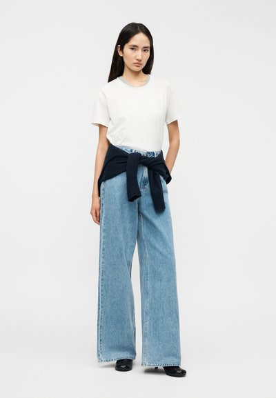 Λευκό κοντομάνικο t-shirt, ανοιχτό μπλε τζιν παντελόνι με φαρδιά παντελόνια και ψηλή μέση, μπλε πουλόβερ δεμένο γύρω από τη μέση και μαύρα μποτάκια μέχρι τον αστράγαλο.