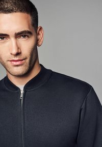 Veste bomber bleu marine foncé avec un col côtelé, une fermeture éclair sur le devant et une texture lisse. Le design présente un style minimaliste et une coupe ajustée.