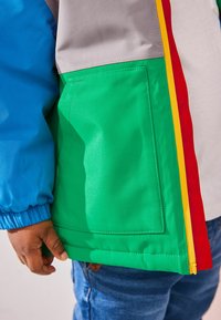 Chaqueta colorida con paneles en azul, verde, gris y rojo. Textura suave con cierre de cremallera y un bolsillo lateral.