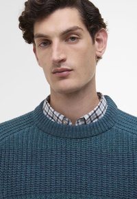 Πλεκτό πουλόβερ σε σκούρο τυρκουάζ, με ριχτό λαιμόκοψη και υφή. Φοριέται πάνω από πουκάμισο με καρό σχέδιο.