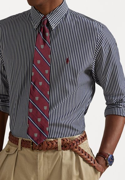 Μπλούζα ναυτικού και λευκών ρίγων, κόκκινη γραβάτα με διαγώνιες μπλε ρίγες και σχέδιο με έμβλημα, χακί παντελόνι, πλεχτή καφέ δερμάτινη ζώνη, καφέ ρολόι.